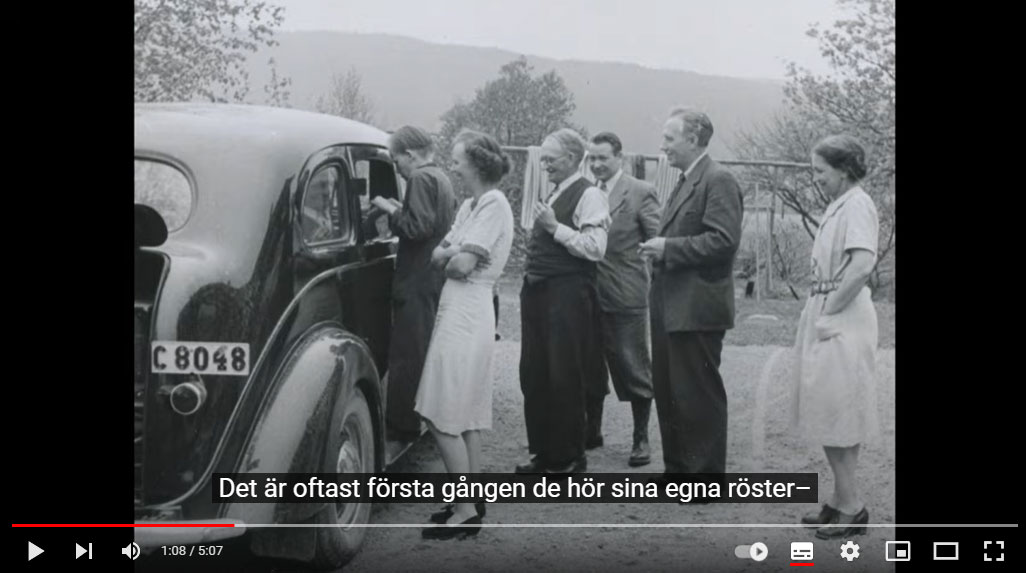 Stillbild från film föreställande en grupp människor som står samlade utanför en bil.
