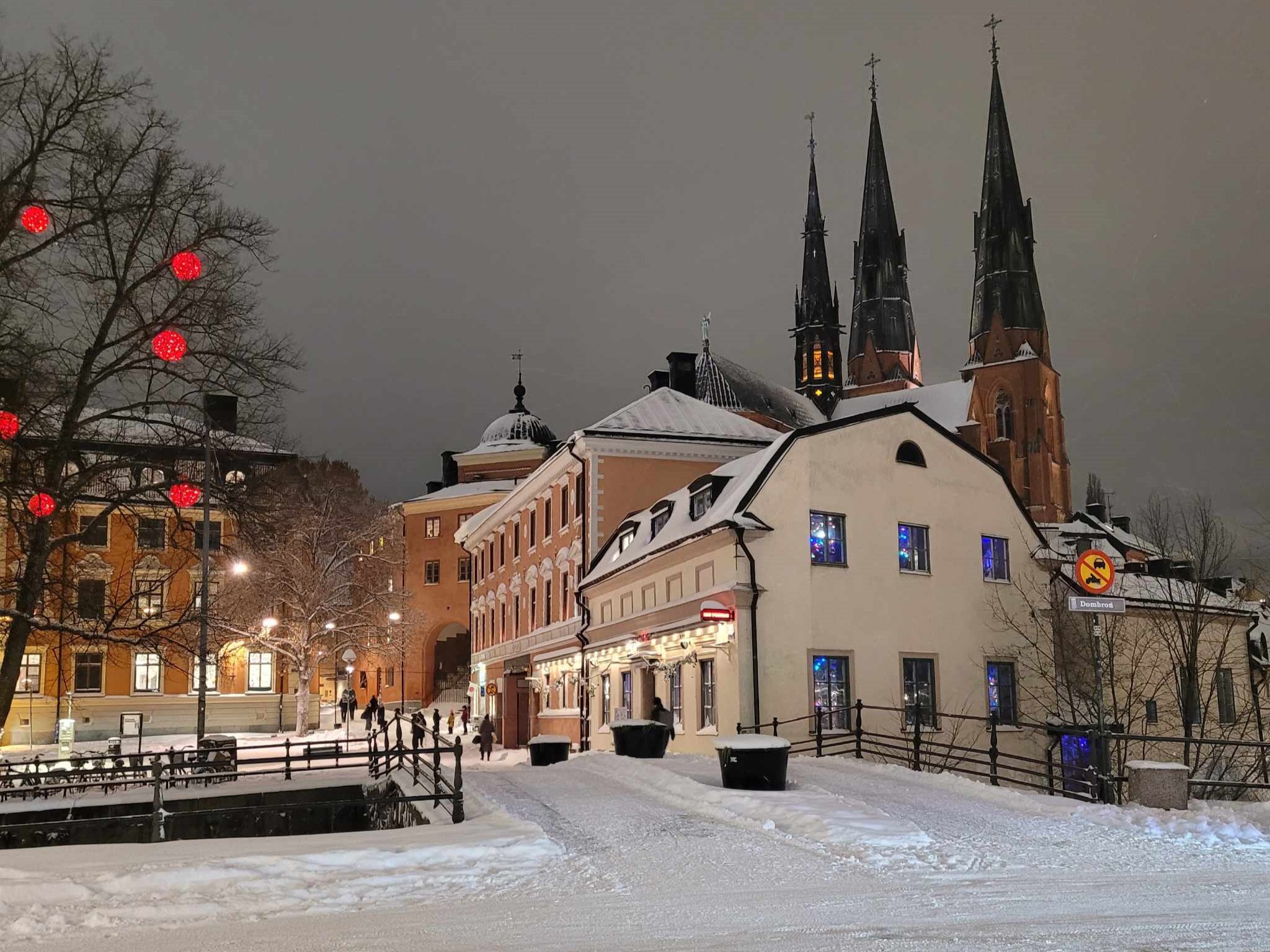 Vy från centrala Uppsala en snöig vinterkväll. I förgrunden syns Dombron och ett träd med belysing av stora röd bollar. I bakgrunden, bakom några hus, syns Uppsala domkyrka. 