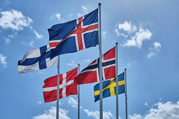 Fem flaggor, från de nordiska länderna, flaggar på stång mot blå himmel.