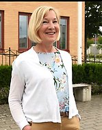 En kvinna med ljust kort hår ler och tittar in i kameran. Hon har en vit kofta och ljusblå topp med blommor på sig.